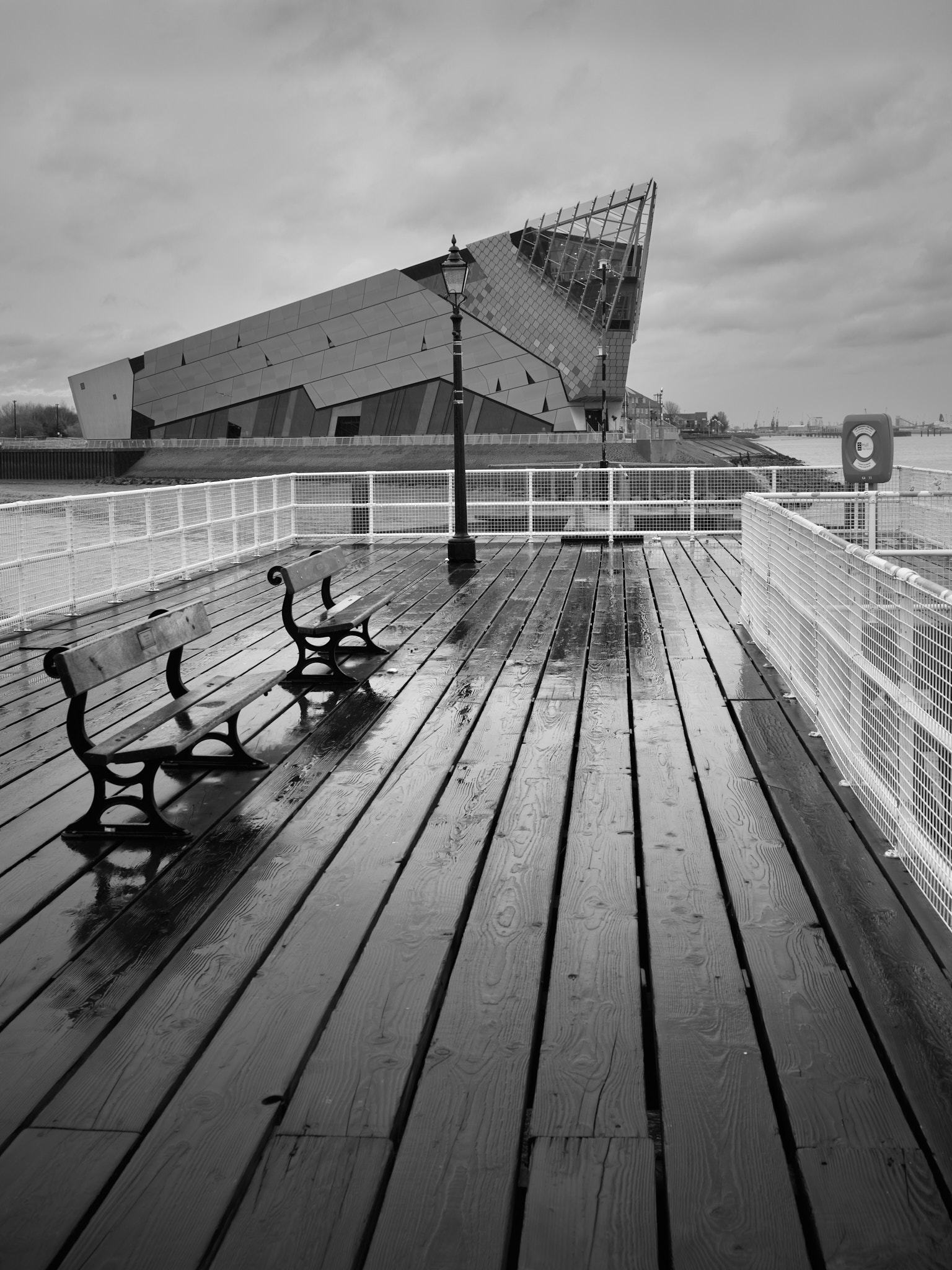 Two wooden benches on the rain-soaked planks of Victoria Pier, a lamp post between them, the angular glass prow of The Deep rising beyond white railings under a grey sky, in black and white