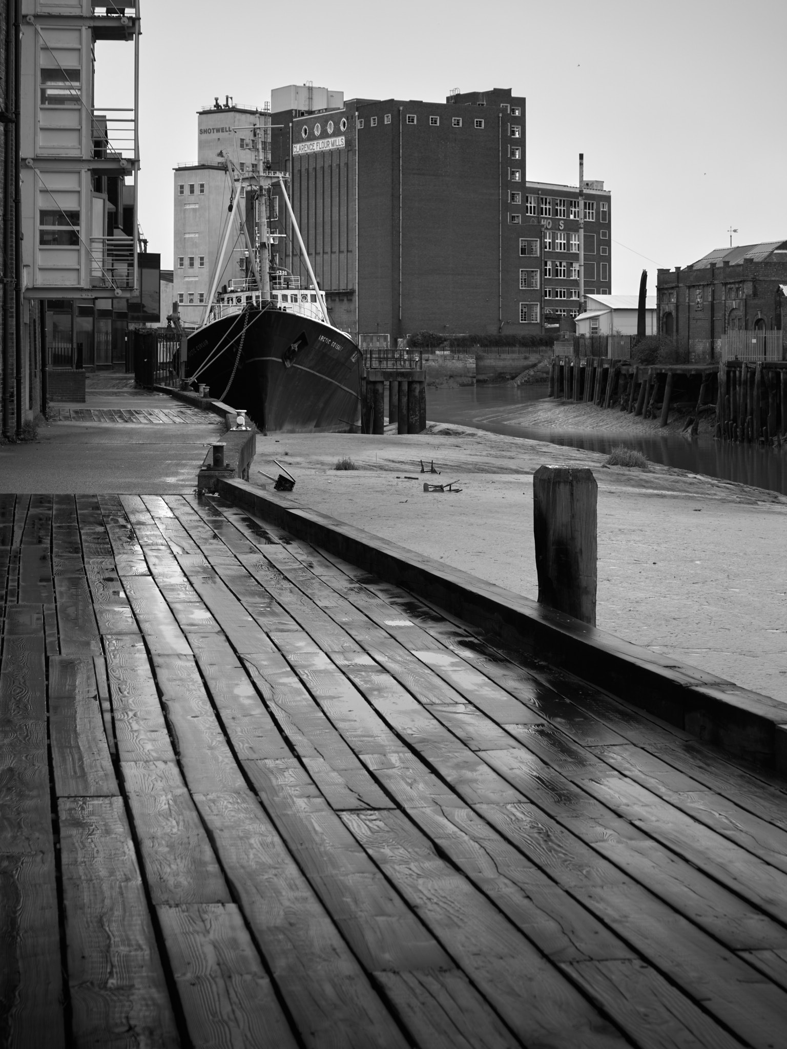 The trawler Arctic Corsair moored on the muddy River Hull beside a wet timber quay, the brick bulk of Clarence Flour Mills behind, in black and white
