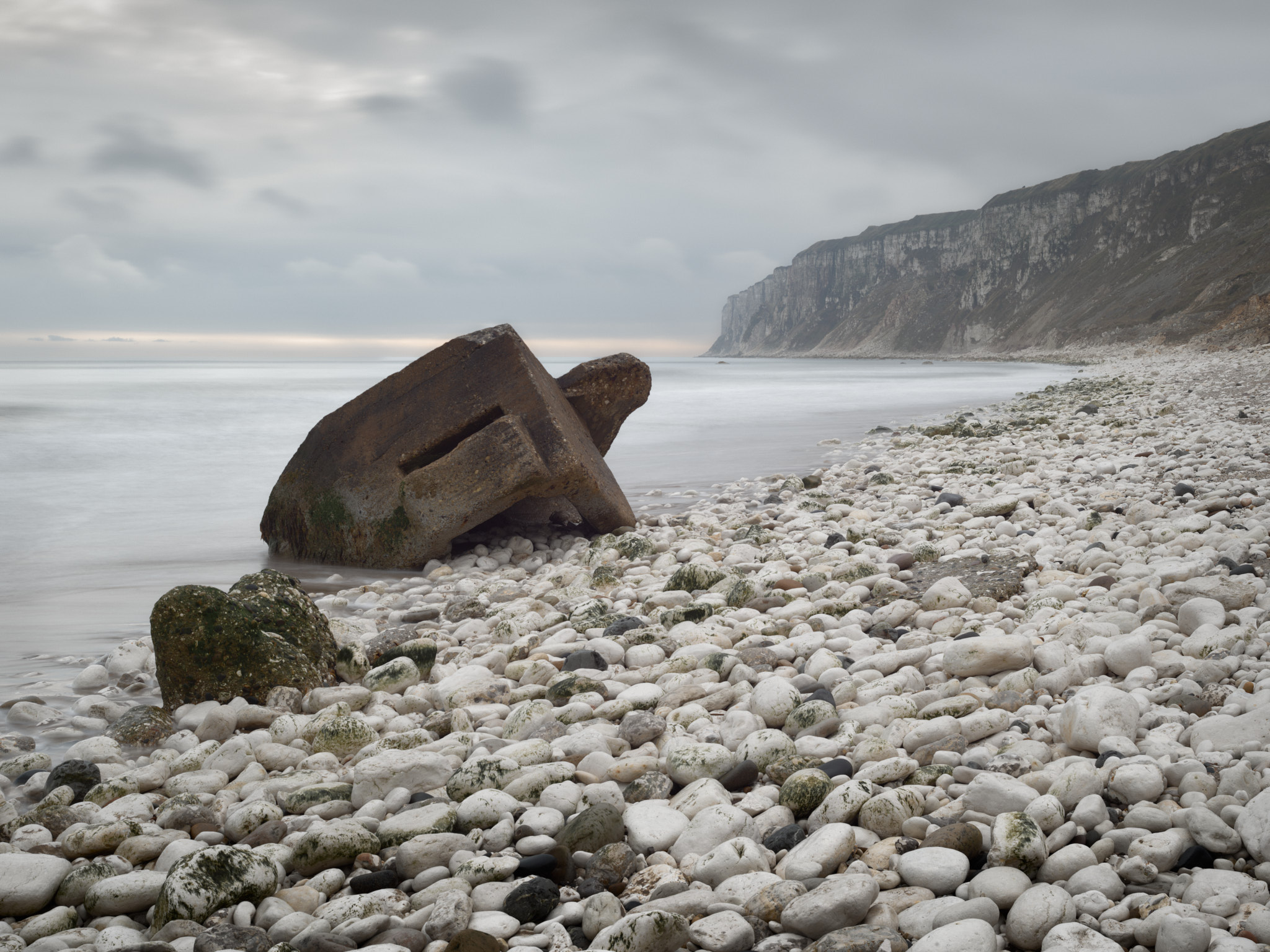 A concrete pillbox tipped on its side at the water’s edge on a beach of white chalk cobbles, the sea smoothed by a long exposure and chalk cliffs fading into grey light beyond