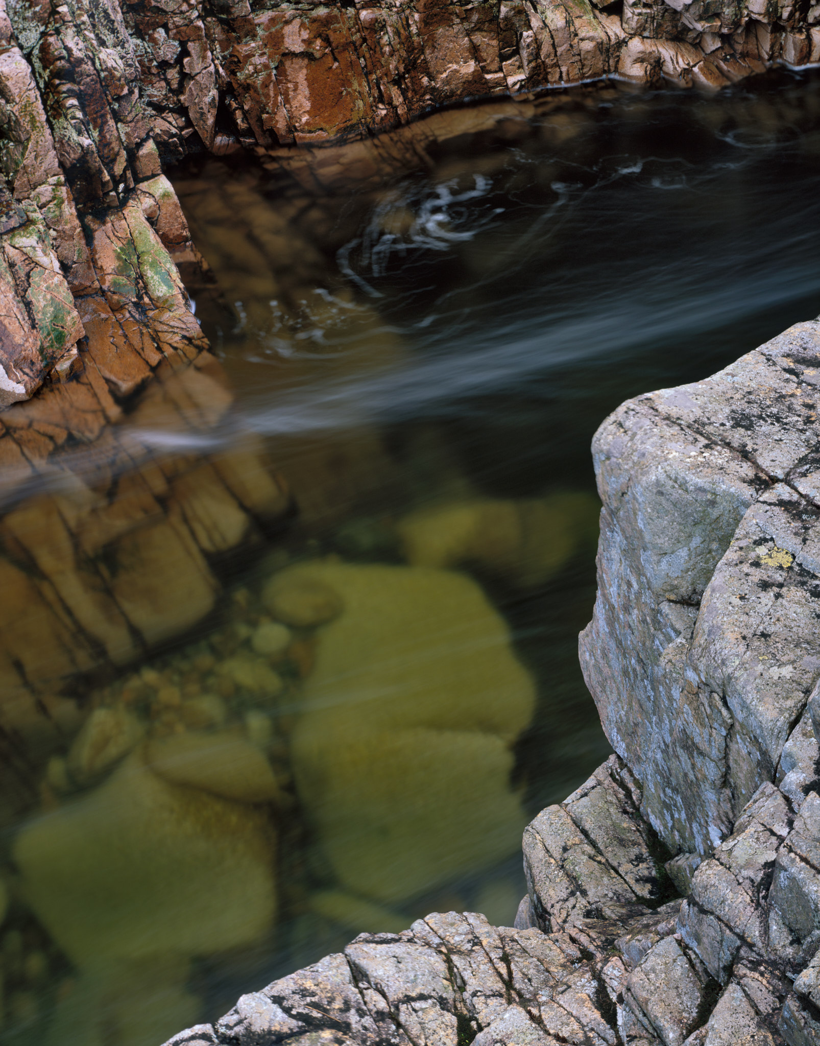 Submerged green boulders beneath the dark, streaked water of the River Etive, red rock above and grey rock in the foreground