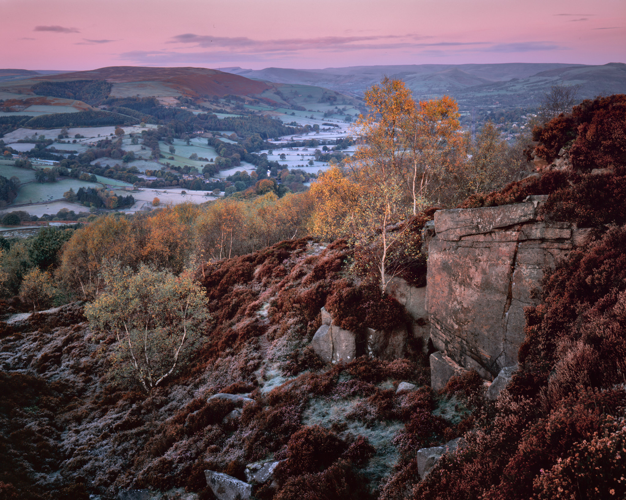 Pre-dawn pink over the Hope Valley from a gritstone edge, frosted fields below and autumn birches on the heather slope