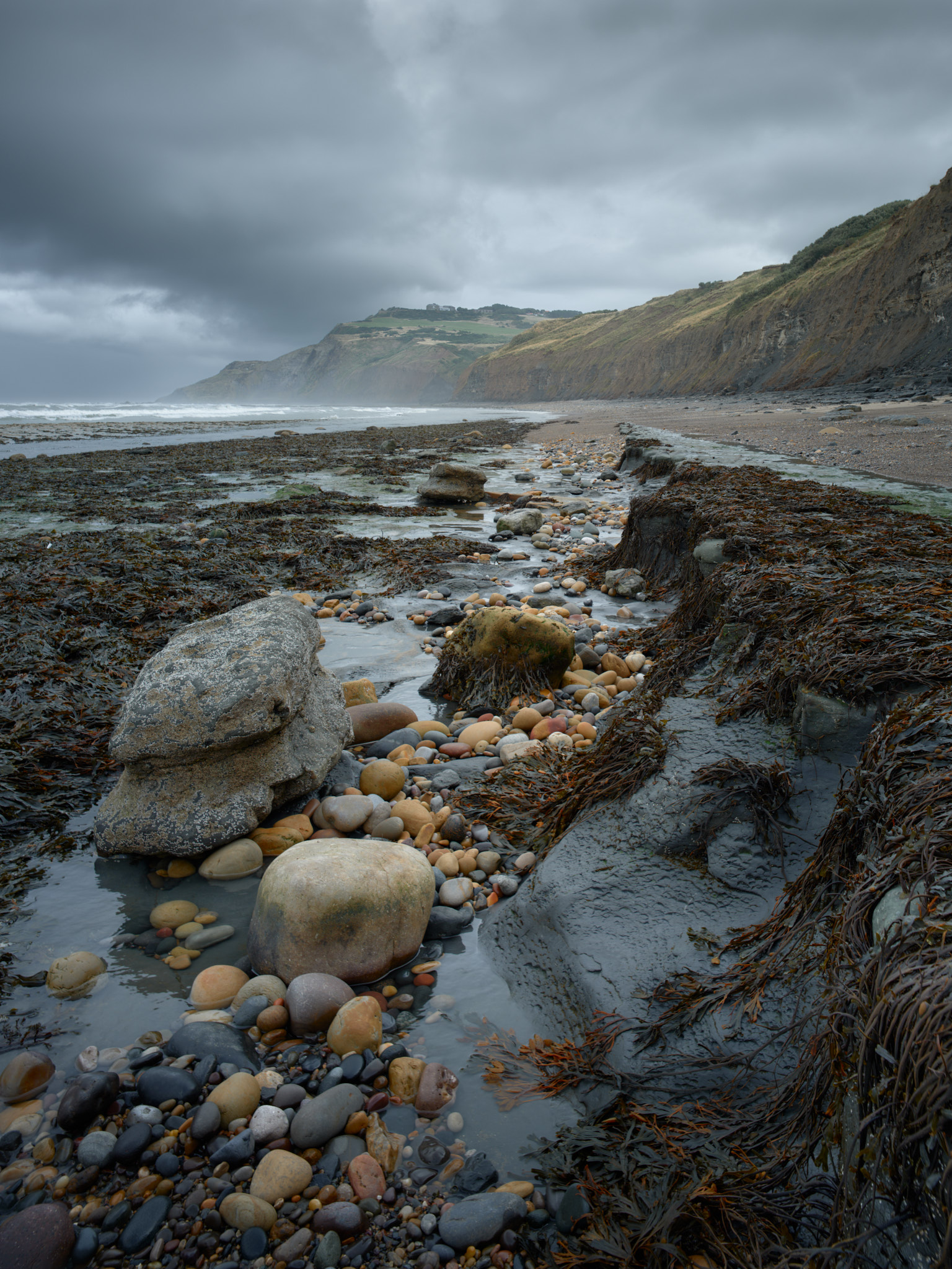 A channel of coloured pebbles and pale boulders between seaweed-draped rock ledges, leading to a headland under a dark storm cloud, with surf breaking in the bay
