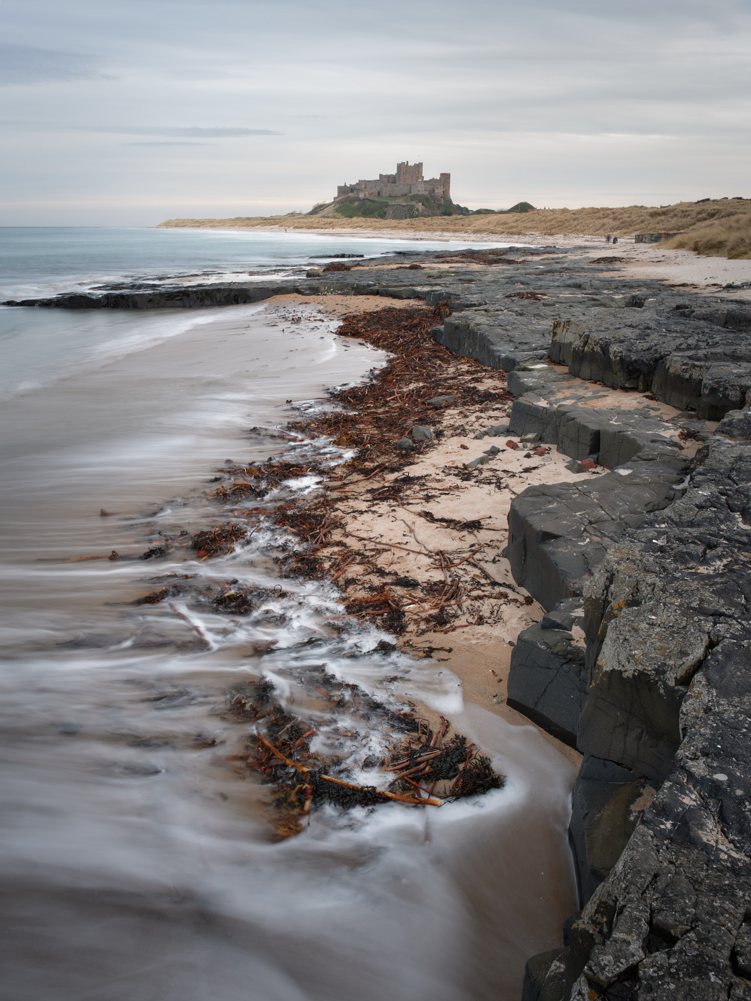 Bamburgh Castle on its crag beyond a curve of beach, a line of red-brown seaweed and dark rock ledges leading towards it, a receding wave blurred by a long exposure