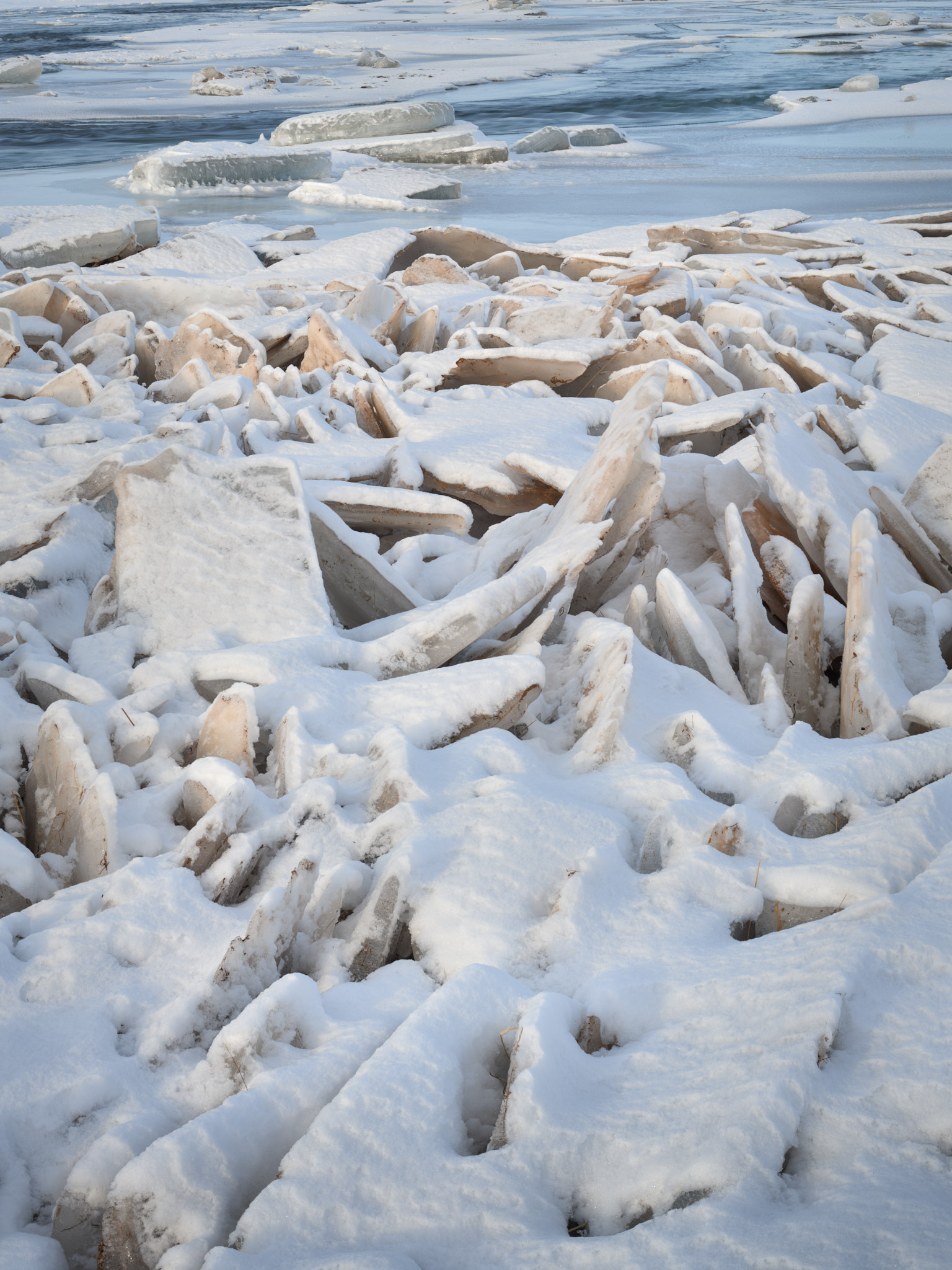A chaos of snow-dusted ice plates tilted and refrozen at the edge of an Icelandic river, open water beyond