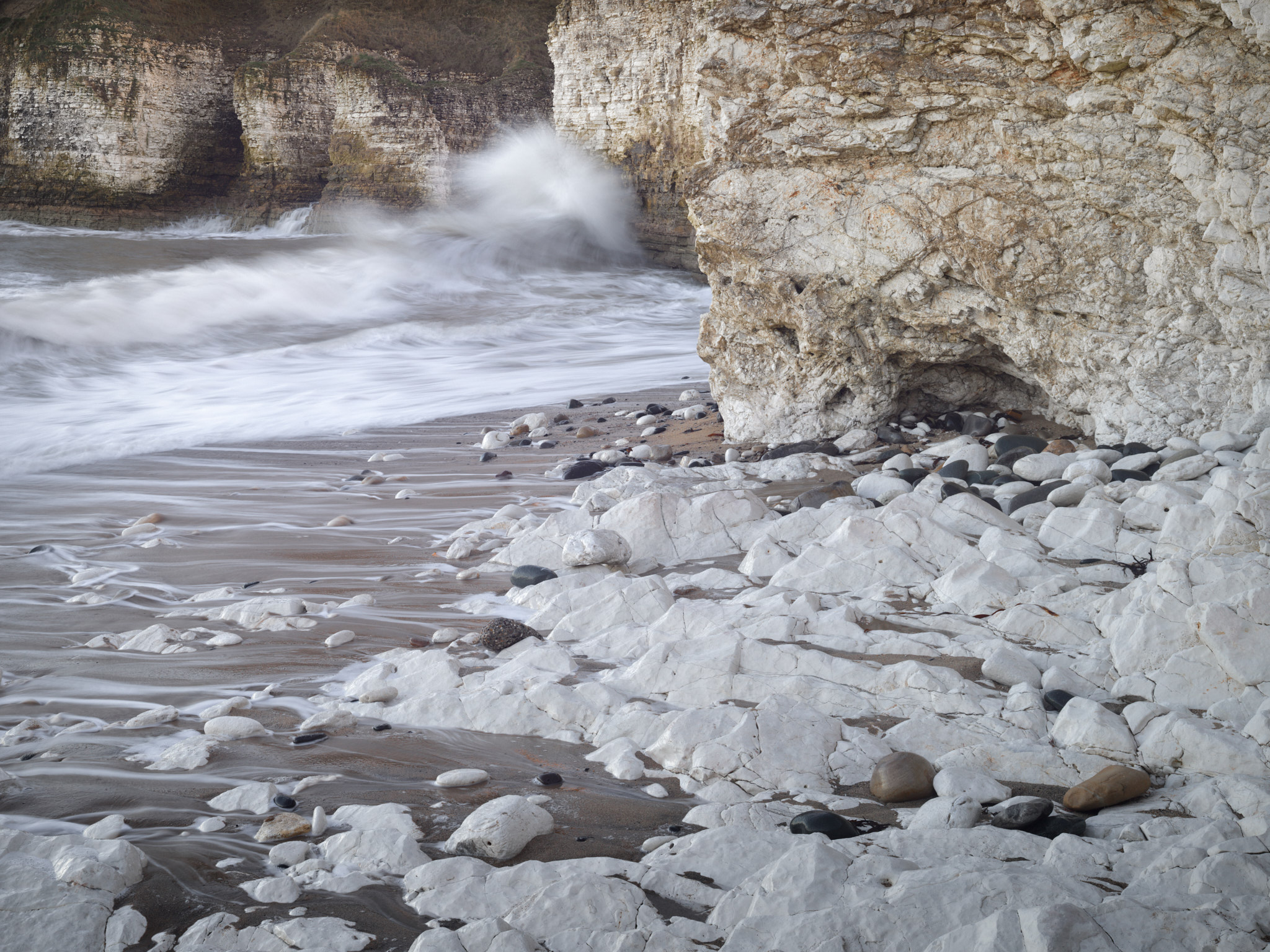 A wave bursting white against the chalk cliffs, wet chalk platform and pebbles in the foreground beneath a buttress of the cliff
