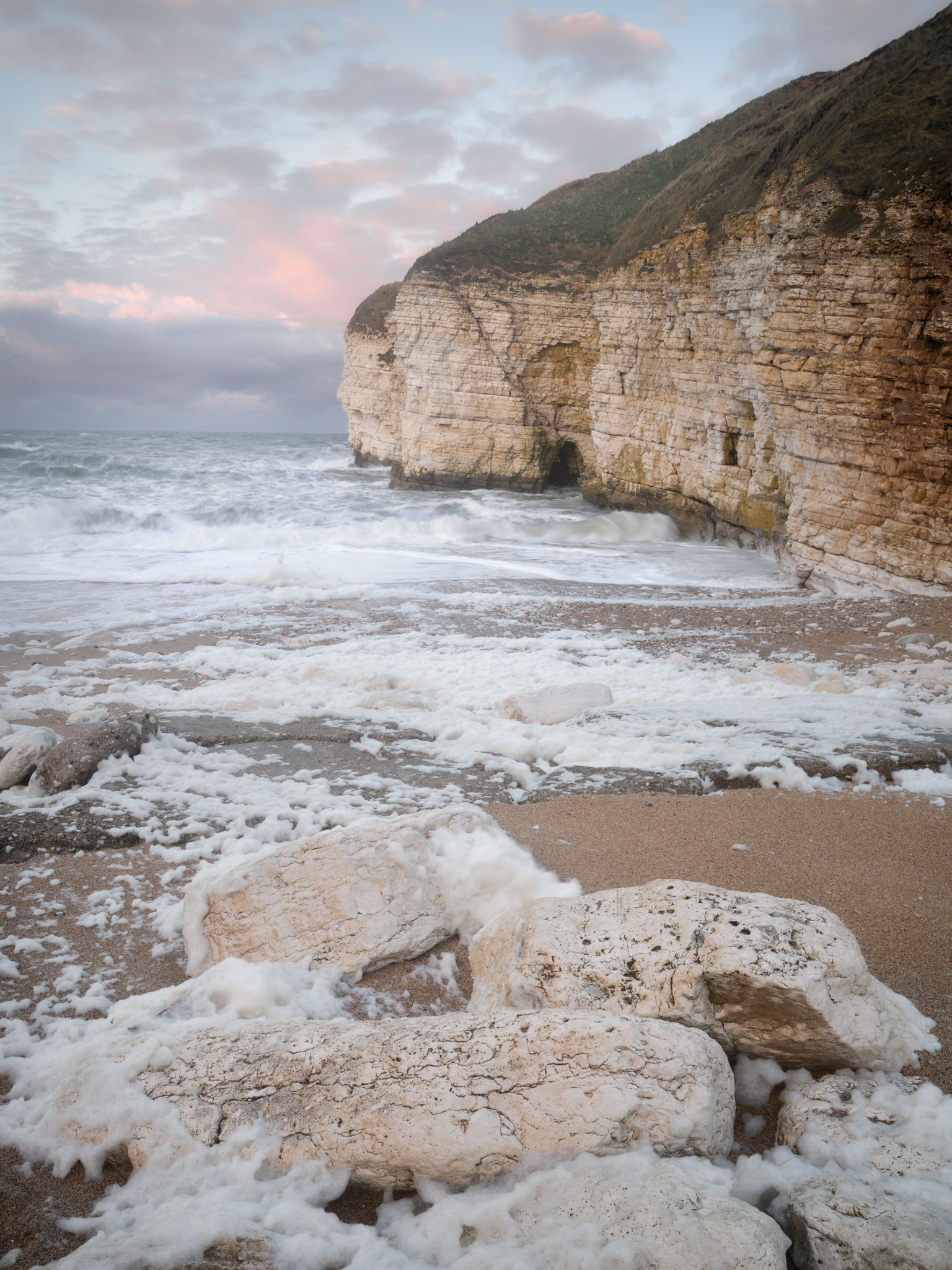 Sea foam heaped over chalk boulders on the beach at Thornwick Bay, a rough sea breaking below the cliffs under a pink-lit sky