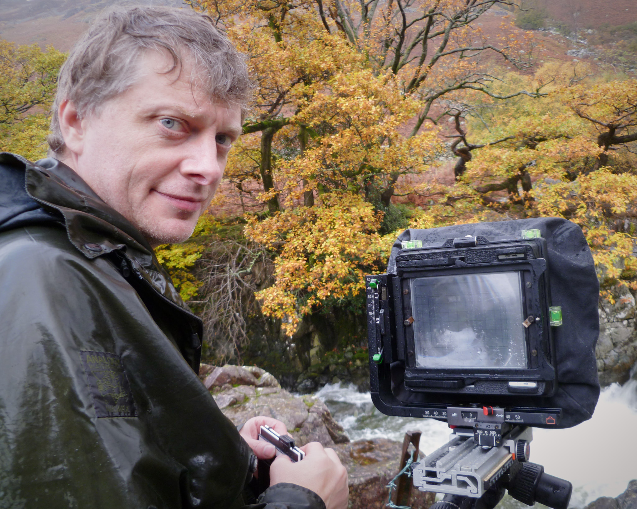 A rain-soaked photographer at a Linhof Technikardan beside a tumbling beck, film holder in hand, autumn oaks behind